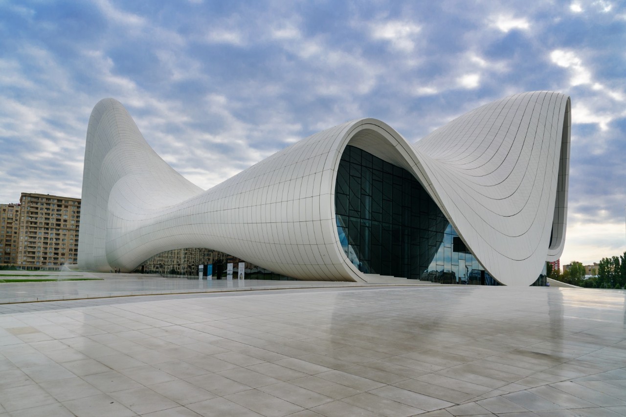 Futuristic, white building with curved shapes and a large window front at the front of the building. The building stands on a large, brightly tiled square. Behind the building, further houses are visible.