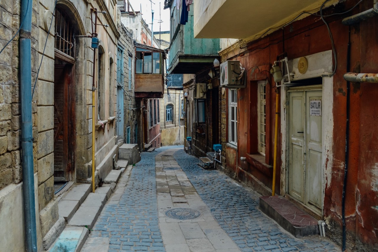 View of a narrow, cobblestone street. Old houses line both sides of the street, with several balconies jutting out over the alley. 