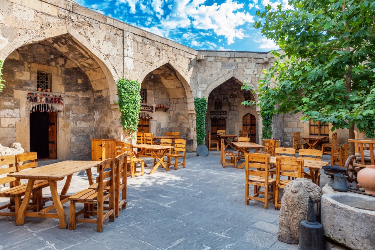 Inner courtyard surrounded by a sandstone-coloured, solid wall, which is interspersed with large, pointed arches set into the wall. There are several tables and chairs in the courtyard. There is a tree on the right side of the picture.