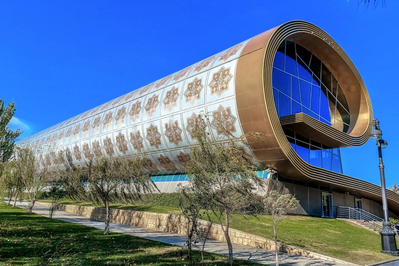 Modern, large building in white with a golden floral pattern, rolled up like a carpet. A window front is visible. There are lawns to the side of the building with a paved path running through them, and a few trees in front of them.