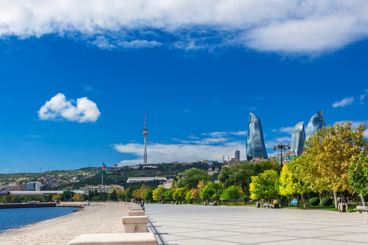 A wide, brightly tiled boulevard, with the sea to the left and trees to the right. The skyline rises in the background. The boulevard is bordered by bollards on the water side.