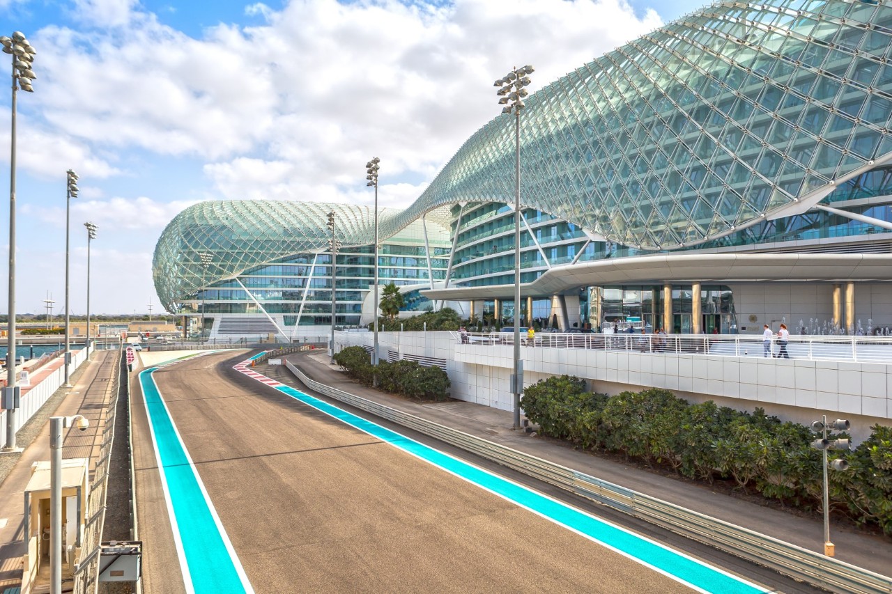The picture shows a modern circuit with a futuristic building on the right. The track has turquoise stripes. The glass building has a curved facade.