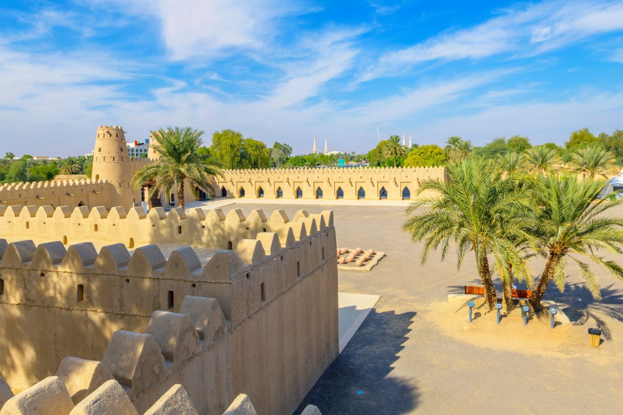 The image shows a historic fortress with sand-coloured walls, a tower and decorations. In the foreground, there are palm trees and a spacious courtyard. 