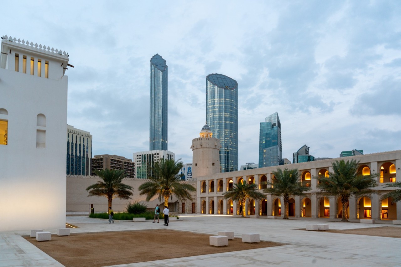 In the foreground of the picture, there is a historic fort with sand-coloured walls and palm trees on a cobbled square. In the background, modern skyscrapers with glass facades are visible. The sky is overcast.