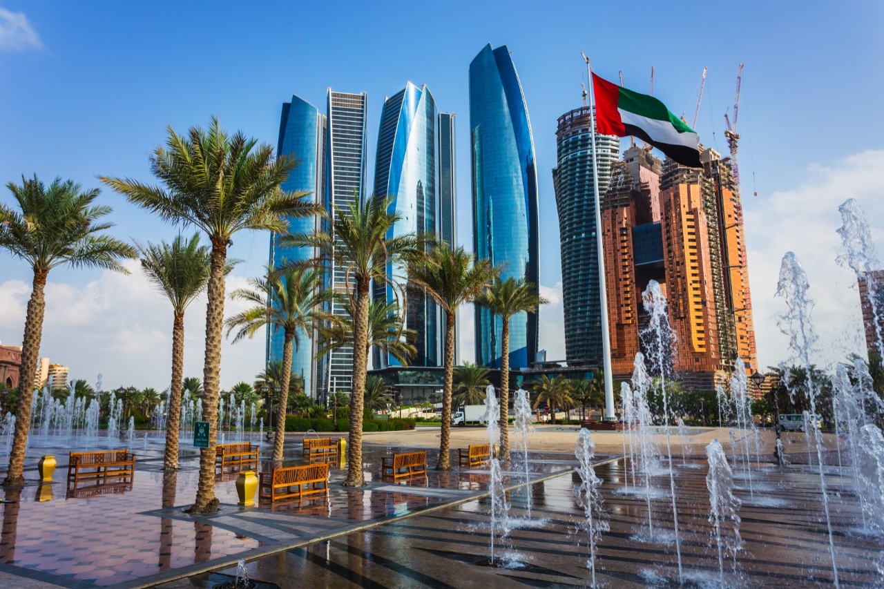 The picture shows a modern skyline with glass skyscrapers. In the foreground, there are palm trees, water features, and benches. A flag of the United Arab Emirates is flying in the wind.