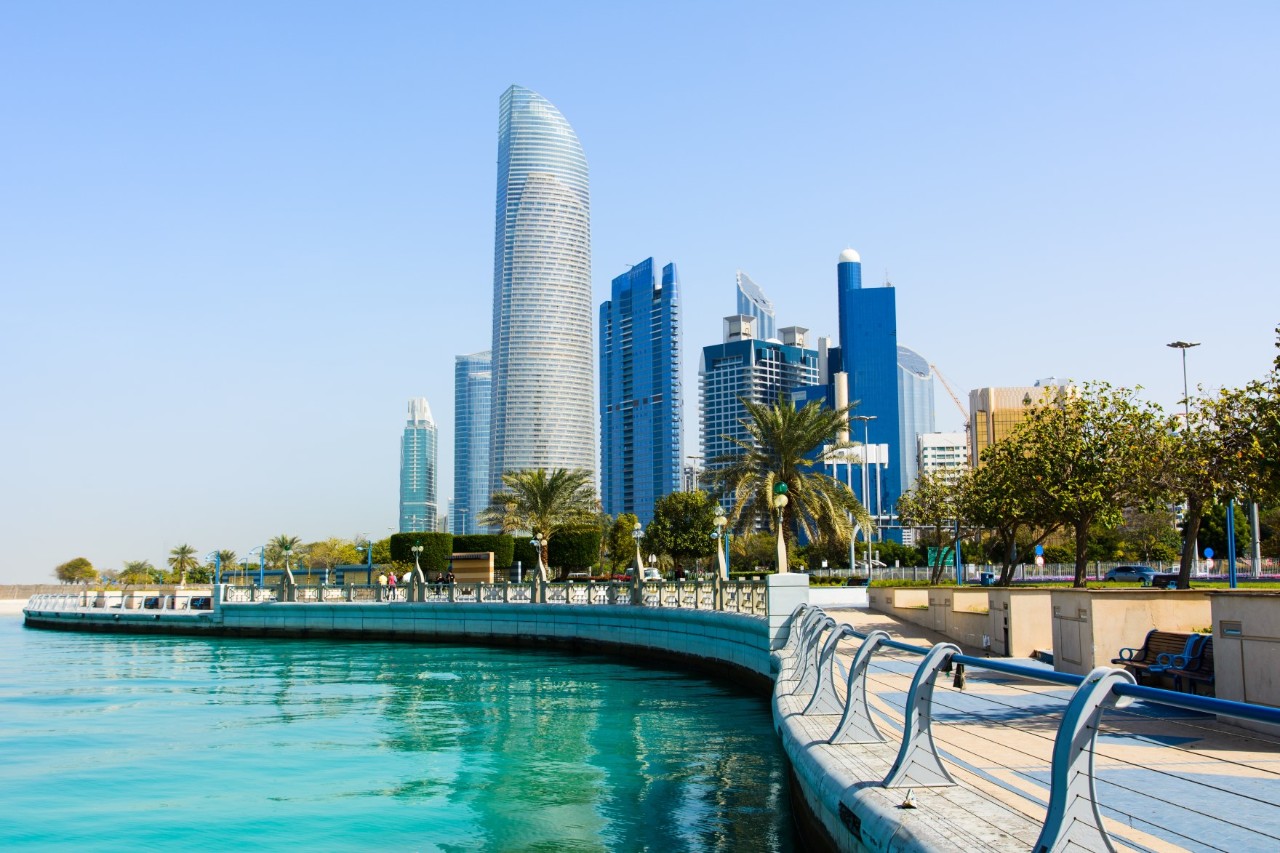 The picture shows a promenade with palm trees and green areas along the turquoise water and several skyscrapers in the background.