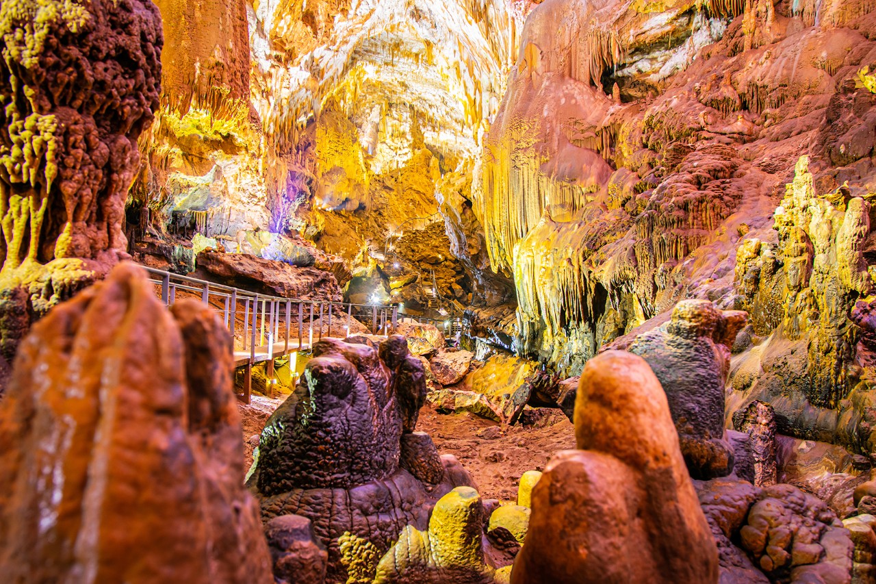 A large, expansive stalactite cave with various rock formations illuminated in warm colors. A small footbridge can be seen on the left side of the picture. 