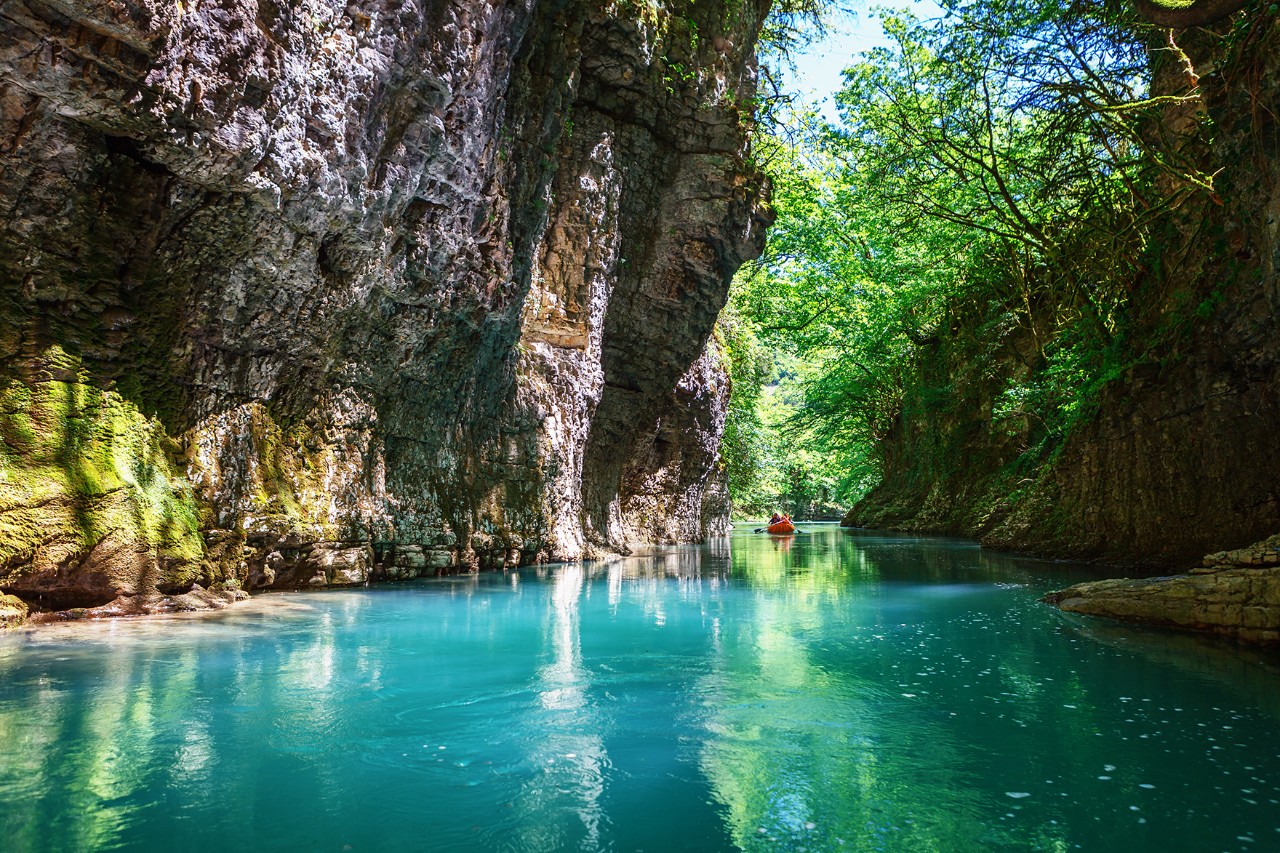 A gorge with steep, moss-covered rock walls and a turquoise blue river. A small red boat and green trees can be seen in the distance.