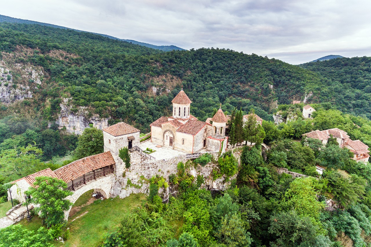 An old monastery on a wooded hill, surrounded by green countryside and a few rocks. The complex consists of several buildings with red tiled roofs and a stone arch.