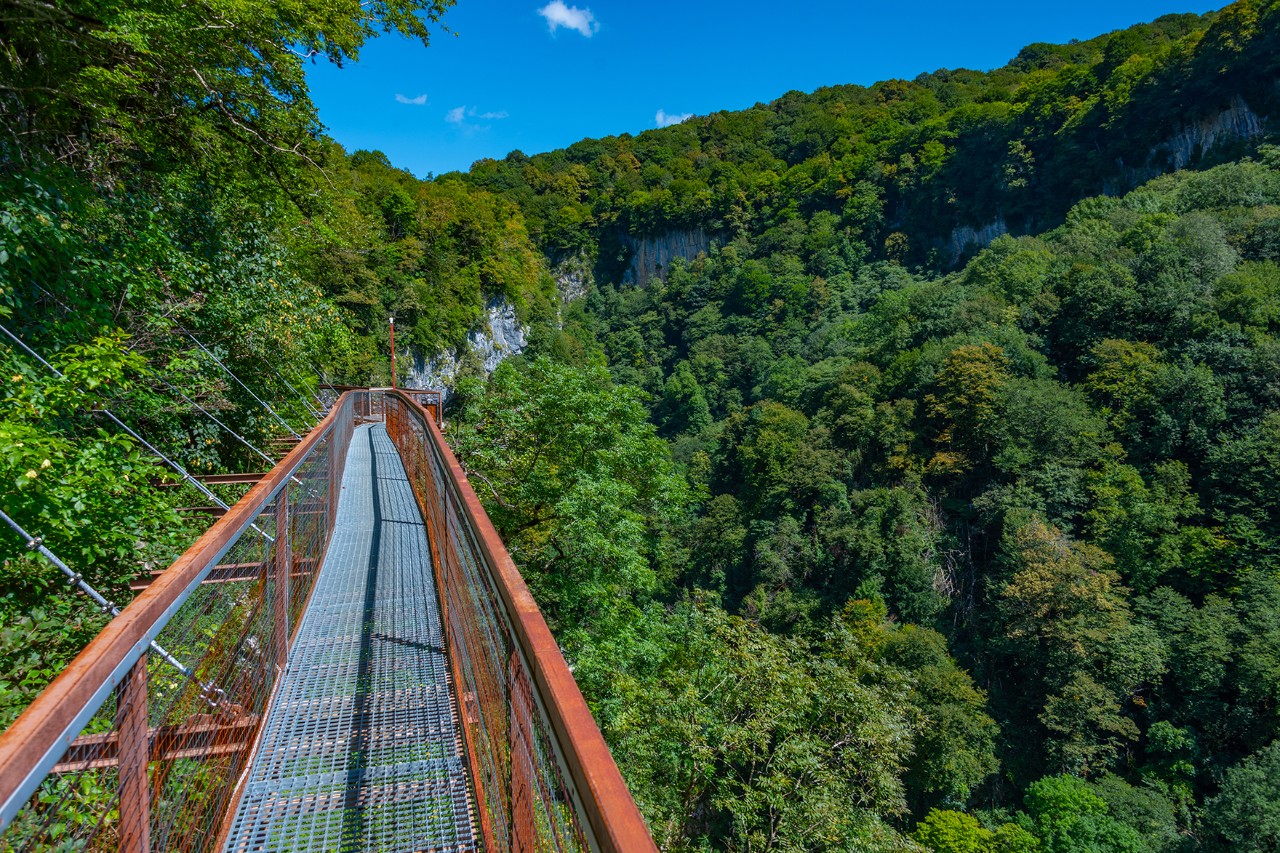 A metal suspension bridge spanning a wooded gorge. The narrow bridge is nestled in lush green surroundings with rock faces.