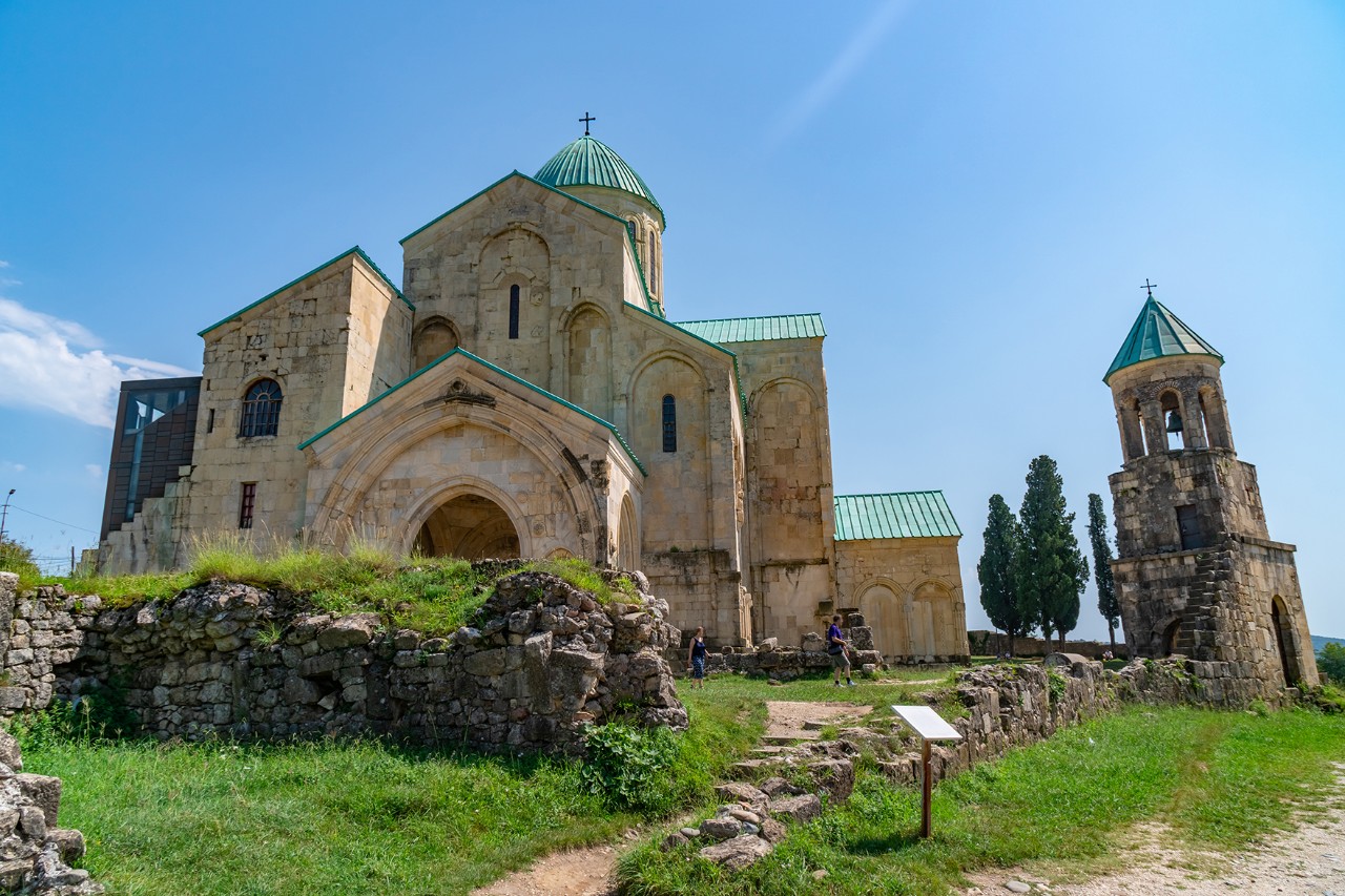 Frog's-eye view of an imposing old church made of light-colored stone with a green roof on a small hill. Next to it stands a separate bell tower. Ruins and a footpath can be seen in the foreground.
