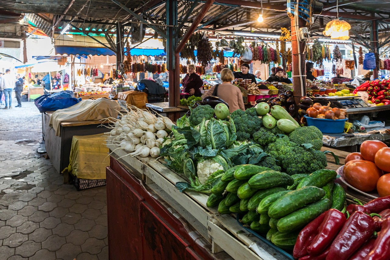 Market stalls with fresh vegetables such as cucumbers, peppers, and cabbage in the foreground. In the background, traders are selling other goods under a covered market stall.