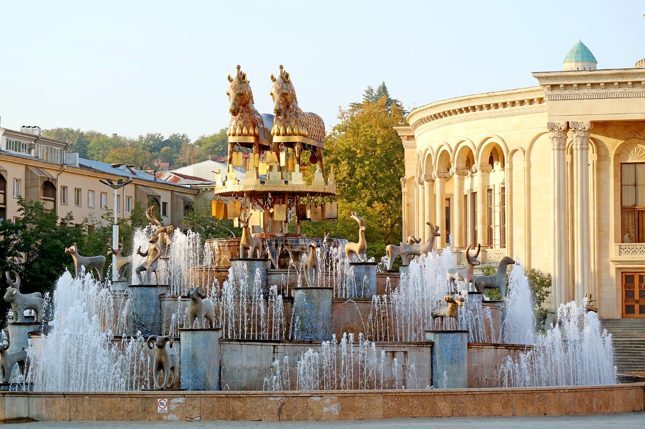 An ornate fountain with golden animal figures and water fountains. Two golden horses stand at the top. In the background is a classic pastel yellow building with columns and round arches.