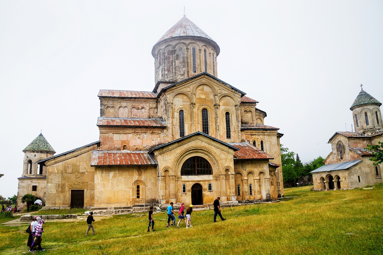 A historic monastery complex with several old buildings. Visitors stroll across a green meadow and explore the grounds.