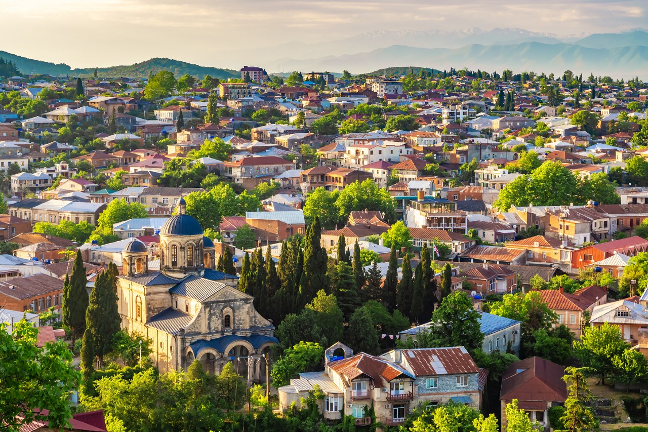 A town with numerous low houses nestled in a hilly, green landscape. In the foreground stands a large, historic church with a dome, surrounded by tall cypress trees.