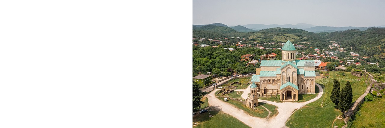 A large cathedral with a light green roof and a dome, surrounded by a green, hilly landscape and scattered houses with red roofs. In front of the cathedral stands a small bell tower. 