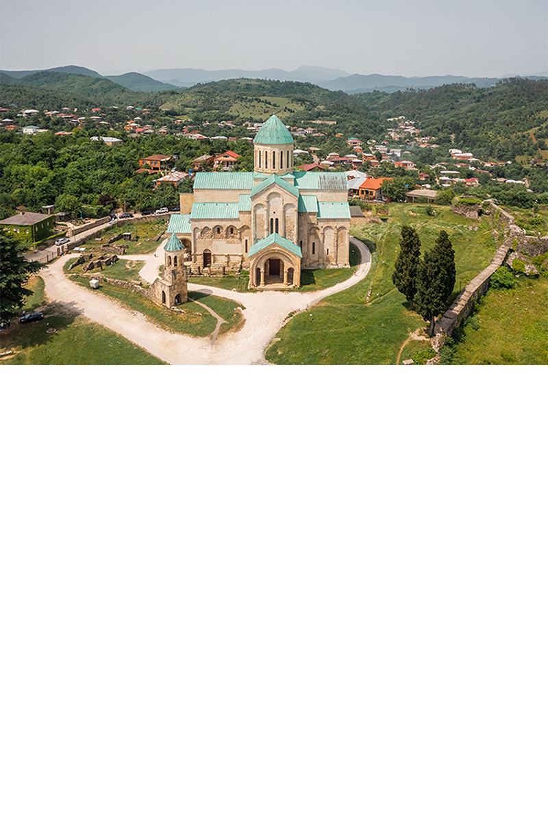 A large cathedral with a light green roof and a dome, surrounded by a green, hilly landscape and scattered houses with red roofs. In front of the cathedral stands a small bell tower. 