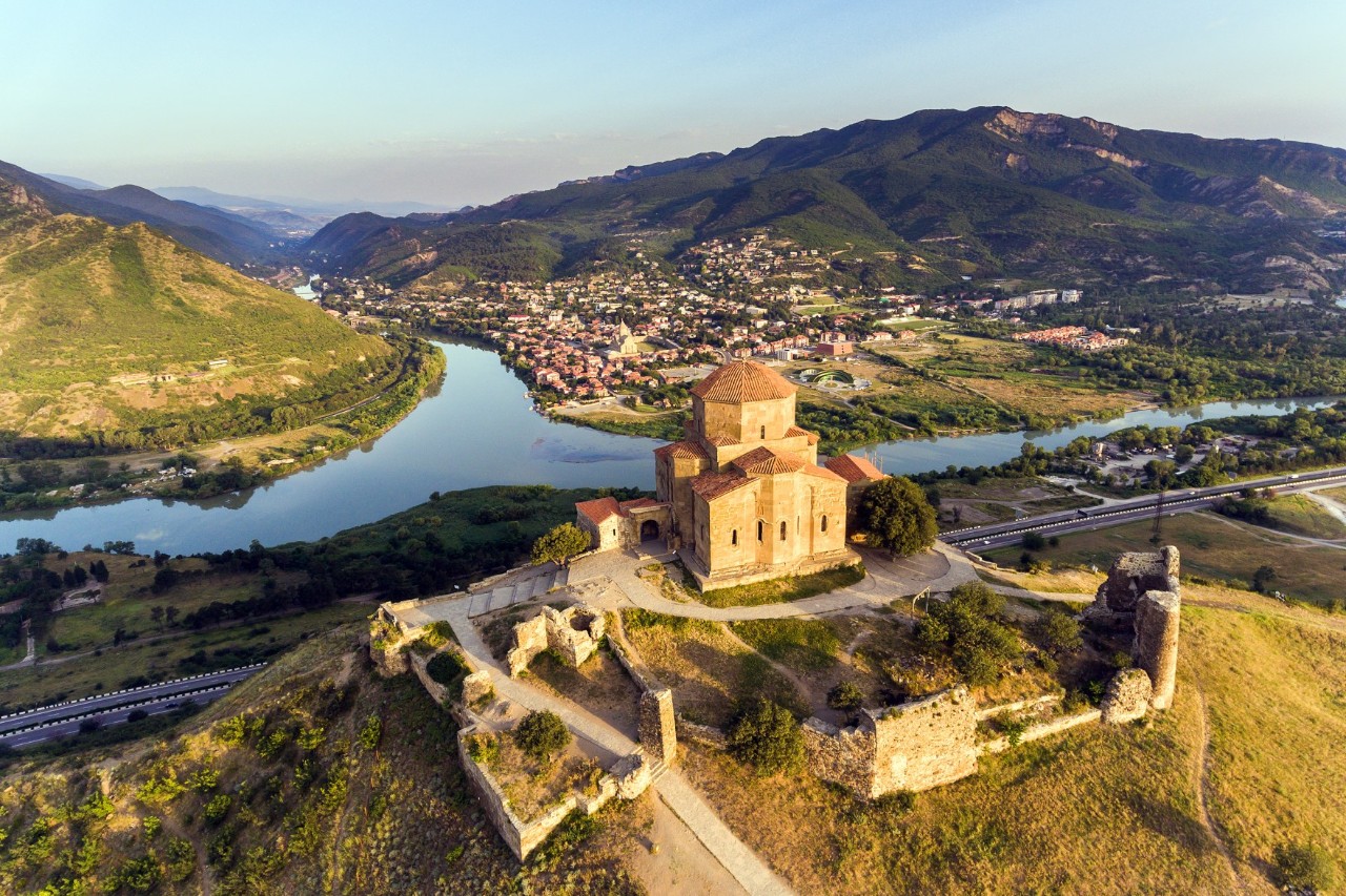 Bird's-eye view of a monastery complex perched on a grassy hill. In the background, a river flows, surrounded by buildings and mountainous terrain.