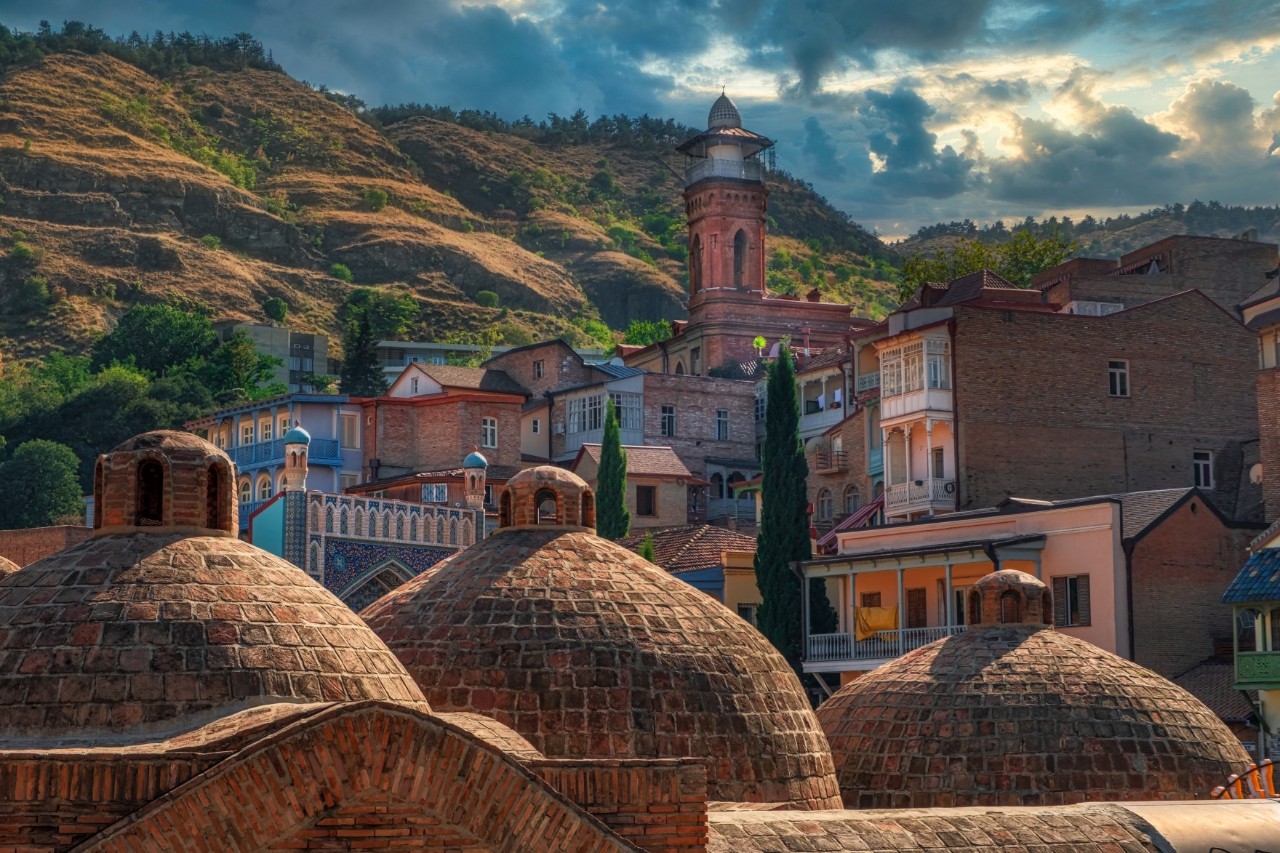 In the foreground, several semi-circular brick domed roofs can be seen. In the background, more houses, green hills and a cloudy sky are visible.
