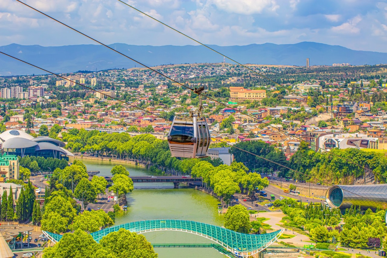 A cable car travelling over a city. Below, a river, city buildings and green spaces can be seen. Mountains stretch into the distance.