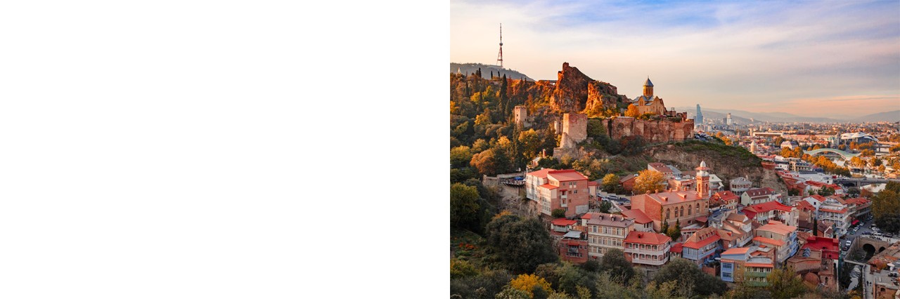 A city in the sunny evening light. In the foreground, there is a densely built-up neighbourhood with red-coloured rooftops. The left half of the picture shows a fortress on a green mountain range. In the background, there is a modern urban area that extends into the mountains.
