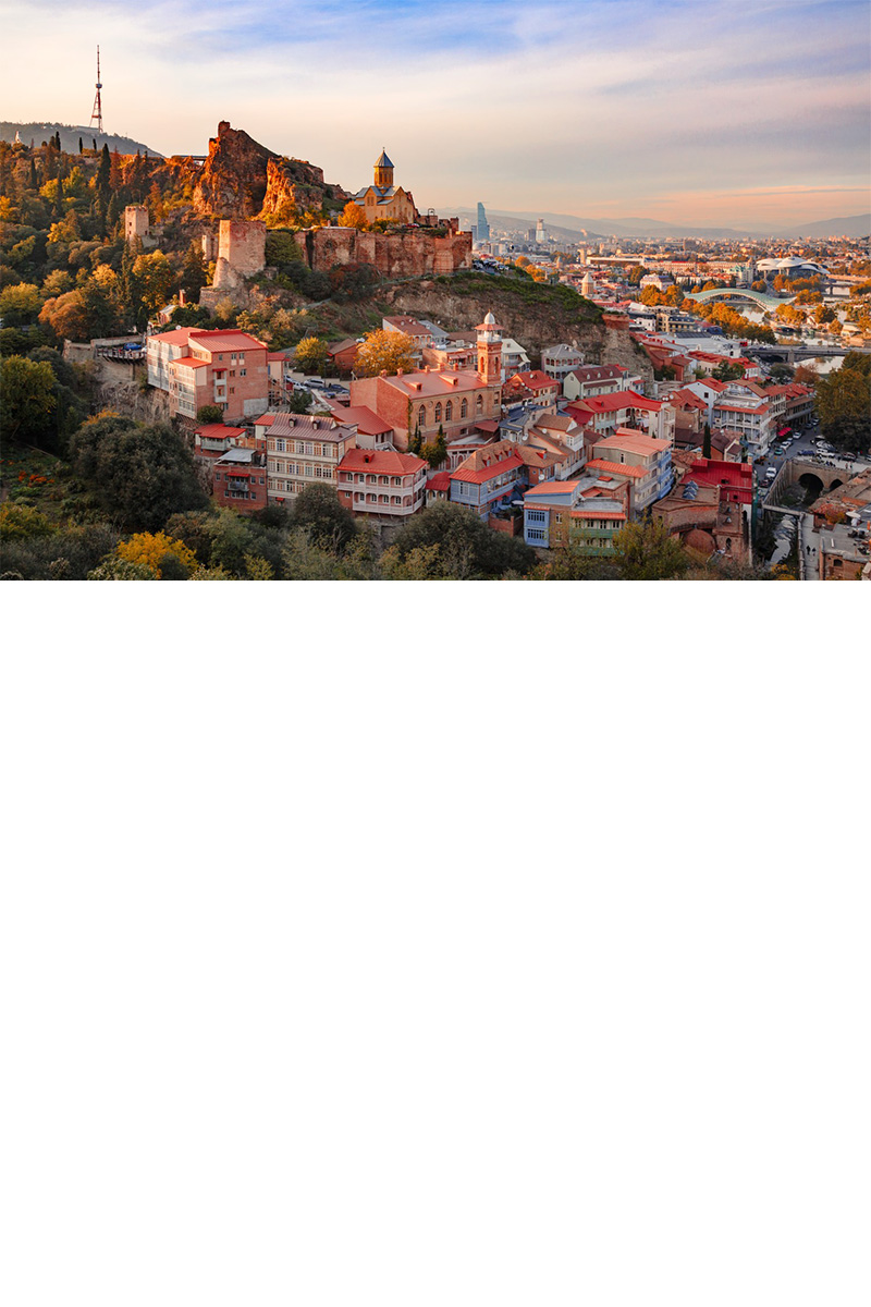 A city in the sunny evening light. In the foreground, there is a densely built-up neighbourhood with red-coloured rooftops. The left half of the picture shows a fortress on a green mountain range. In the background, there is a modern urban area that extends into the mountains.