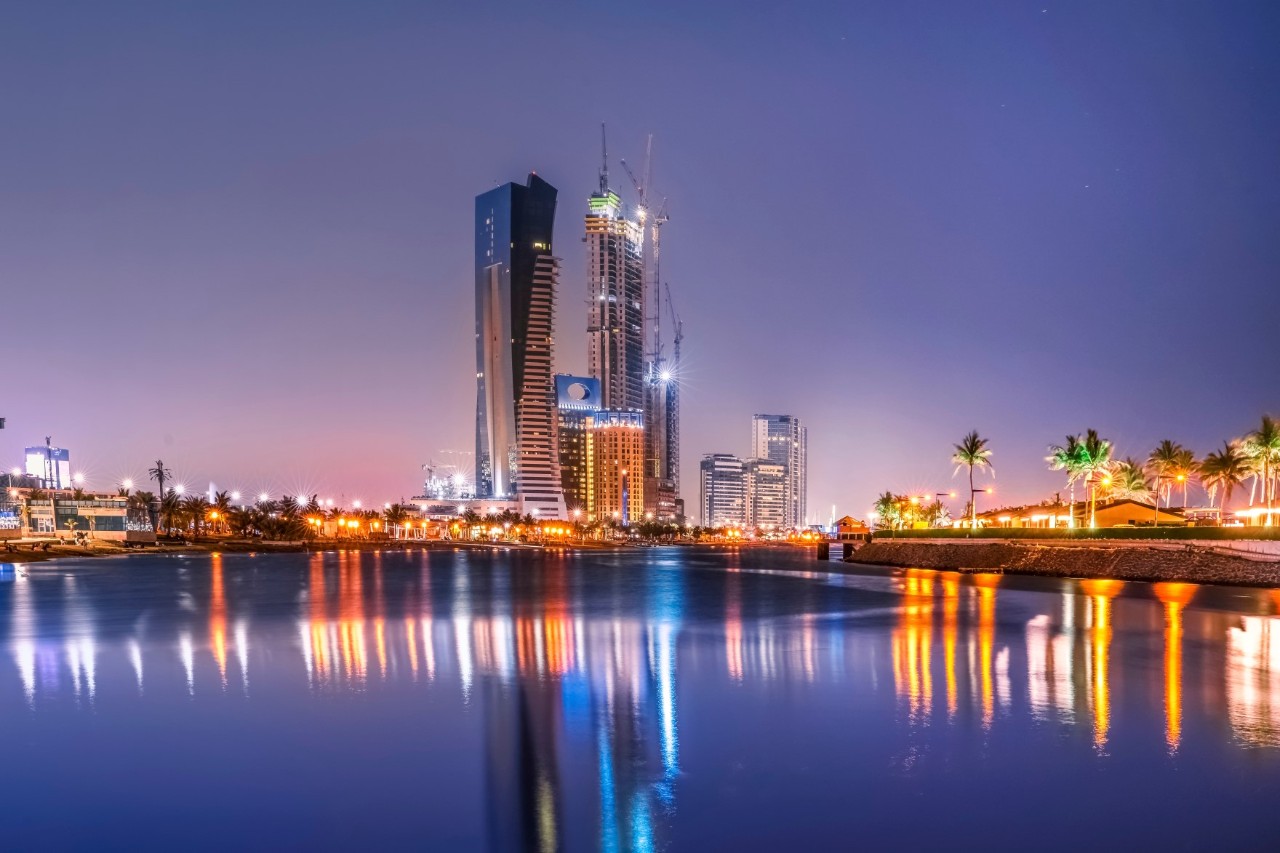 The coast from the water, evening atmosphere with illuminated skyscrapers, smaller buildings and palm trees. 
