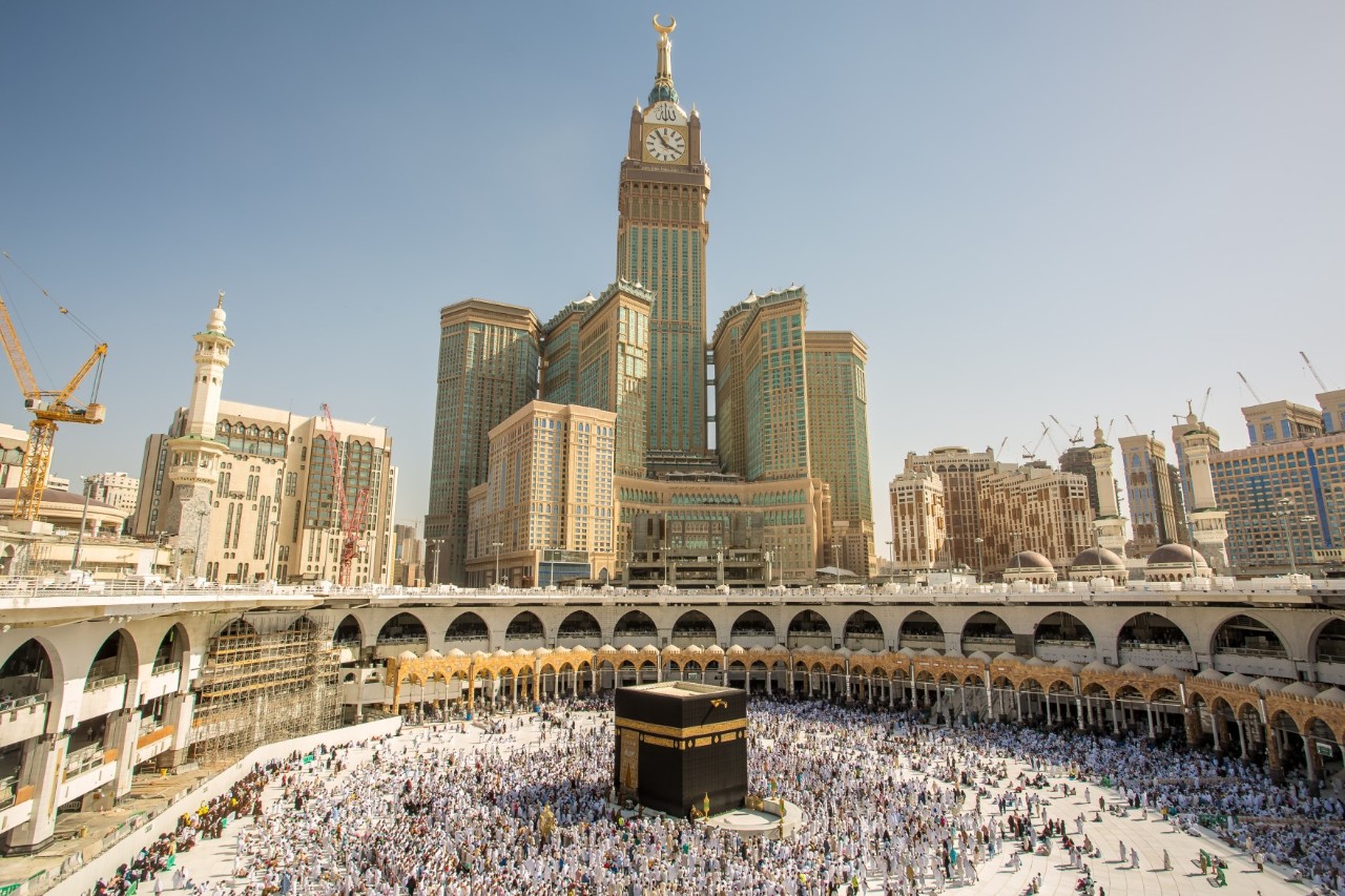 Inner courtyard of the Holy Mosque with many people around the black, cuboid Kaaba surrounded by skyscrapers, hotels and bell tower.