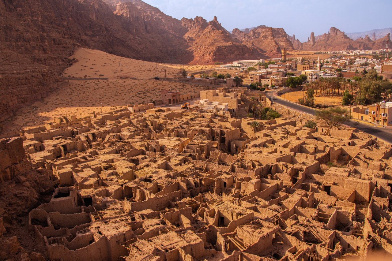 An excavation site with sand-coloured houses and rock tombs surrounded by mountains and a village in the background. 