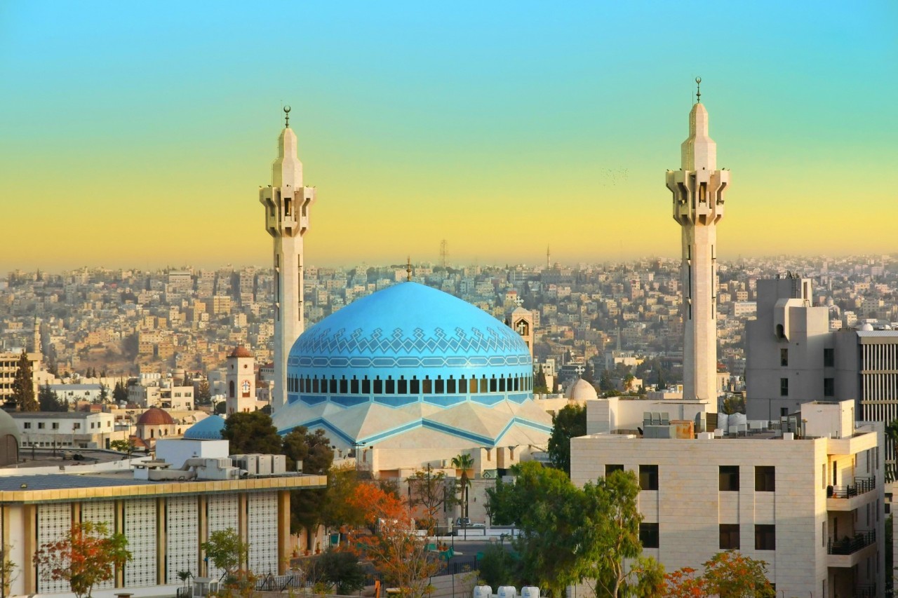 View of a very large mosque with a blue dome and two towering minarets. In the foreground are large, light-coloured buildings. In the background a densely built-up sea of houses.