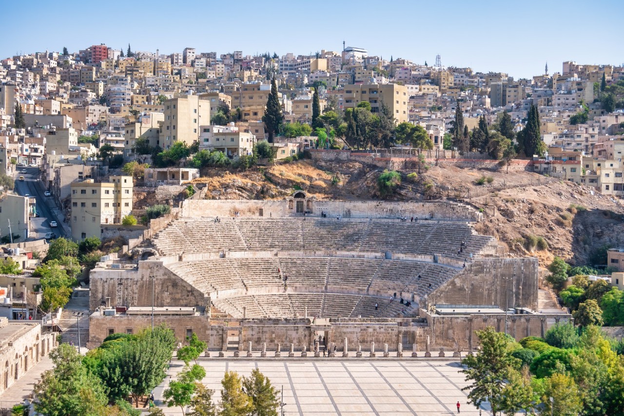 View of a semi-circular amphitheatre with three tiers located directly on the hill. Behind it is the sea of houses in the city centre. In front of it is a deserted square surrounded by green trees.