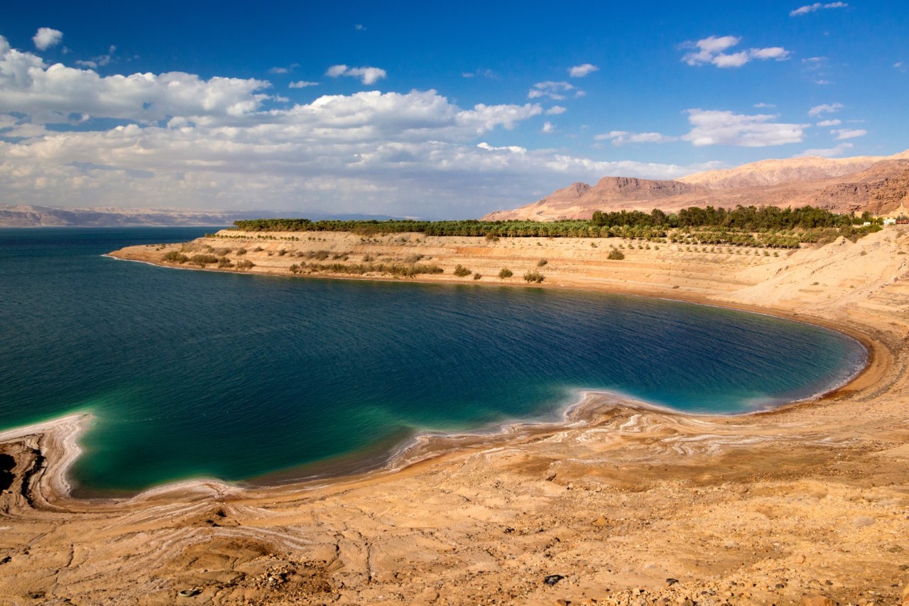View of a turquoise-blue lake surrounded by a barren, rocky, desert-like shore with hills. Salty deposits can be seen on the shore. On the right of the picture, a high headland protrudes into the water with trees growing on it.
