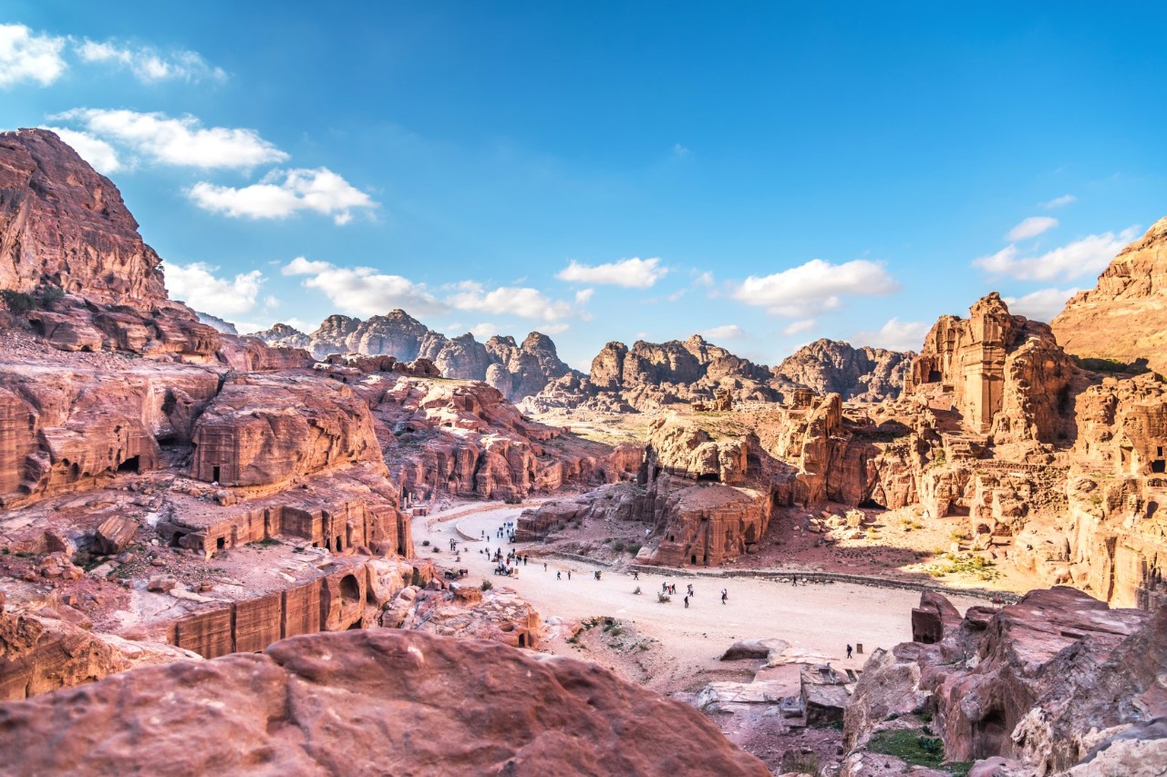 View of an extensive rock city with a wide path running through it. Stone-carved facades can be seen in some of the rocks. The part of the town on the left of the picture is in the shade, the rocks appear pink in colour. The part on the right is illuminated by the sun, the rocks appear yellow to brownish in colour. The sky is blue and slightly cloudy.
