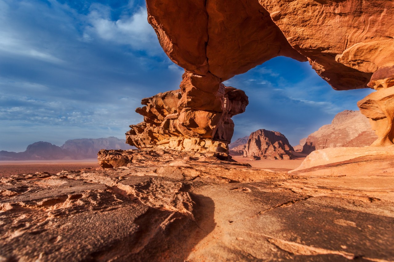 View of a bizarrely shaped rock formation made of red sandstone, which is part of a rocky desert landscape with mountains. The rock gives the appearance of a kind of arch, creating an opening.