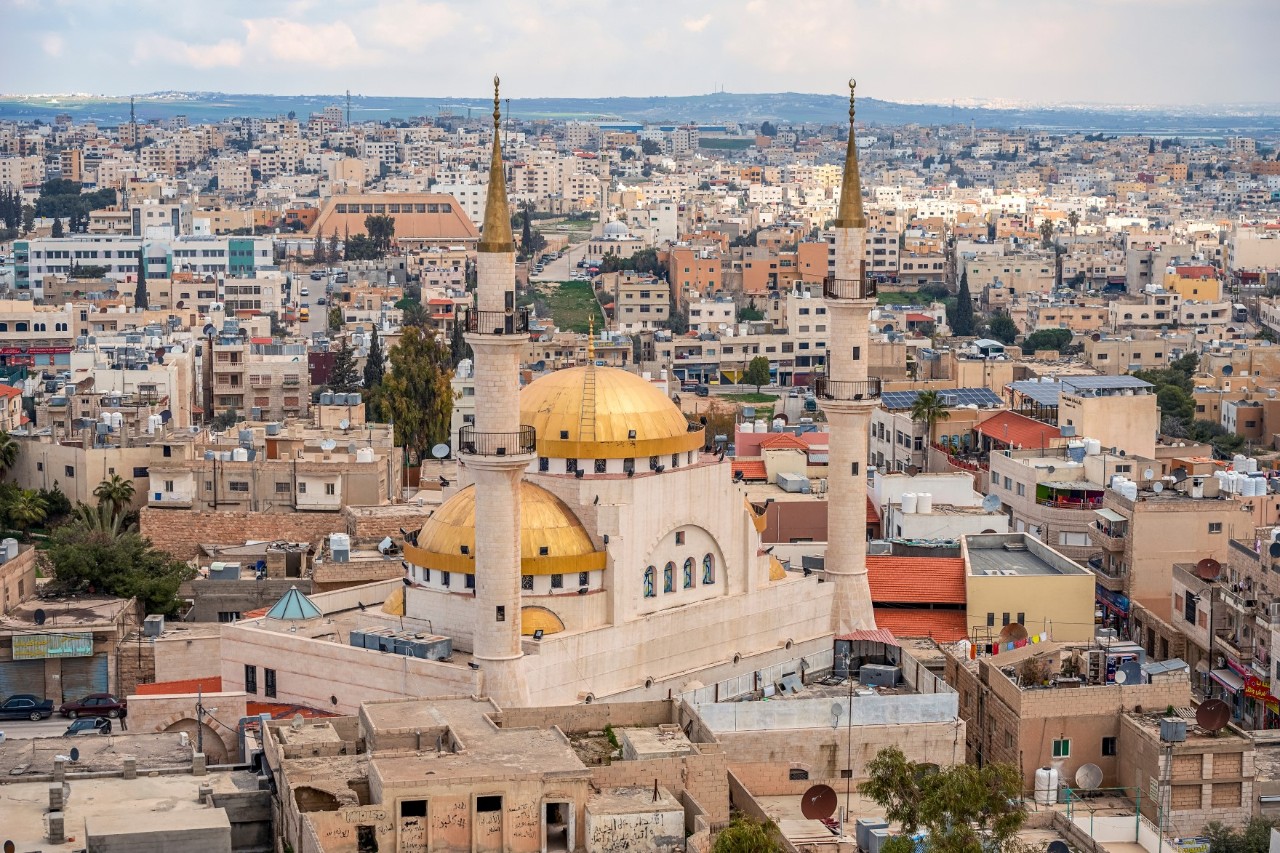 View from above a mosque with two golden yellow domes and two minarets in the centre of a densely built-up city. The sea of houses continues to the horizon.