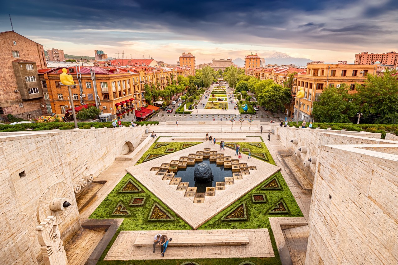 View from the steps of the sandstone-coloured Cascade monument to an adjacent boulevard flanked by reddish buildings and trees. Within the Cascade is a work of art on the ground with ornaments, water basin and a sphere.