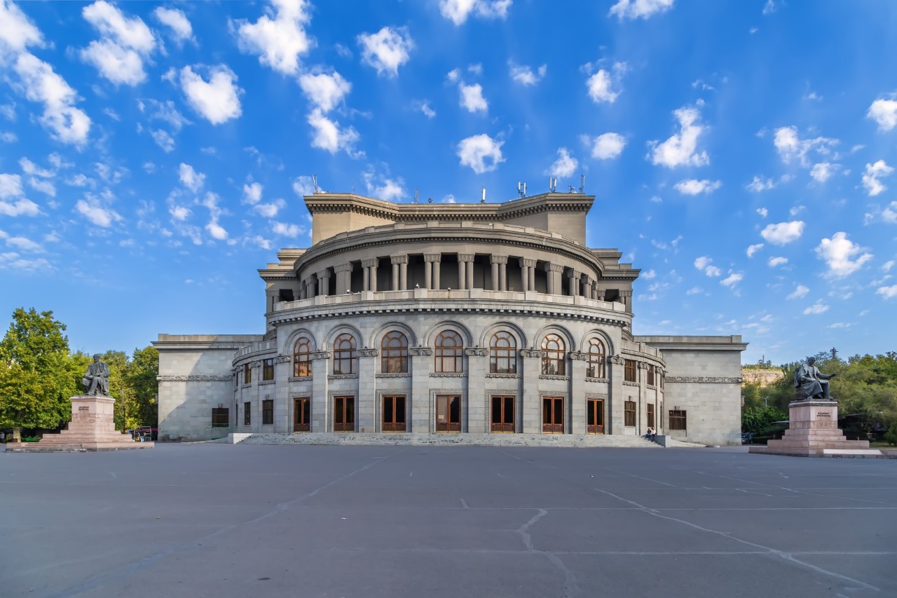 View of an impressive neoclassical-style building with a wide, columned vestibule with large arches, semi-circular balcony and curved dome. To the left and right of the building is a massive sculpture and trees, and in front of the building is a large, deserted square. The sky is blue and slightly cloudy.