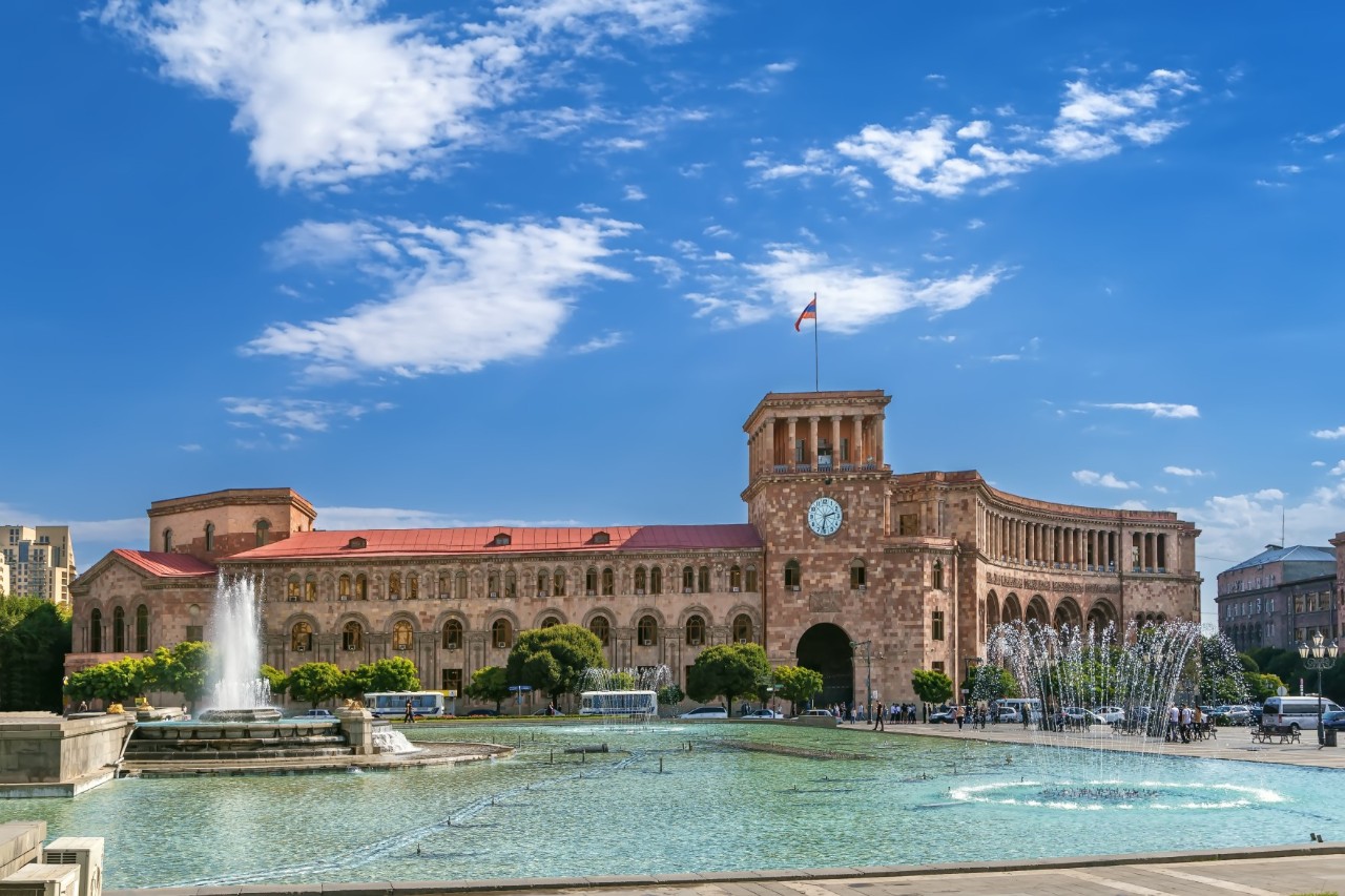 View of an elongated, grand building with many arched windows and a large passageway. There is a clock on the rectangular tower of the building and a flag on the tower. A large water basin with a fountain and a spring can be seen in front of the building.