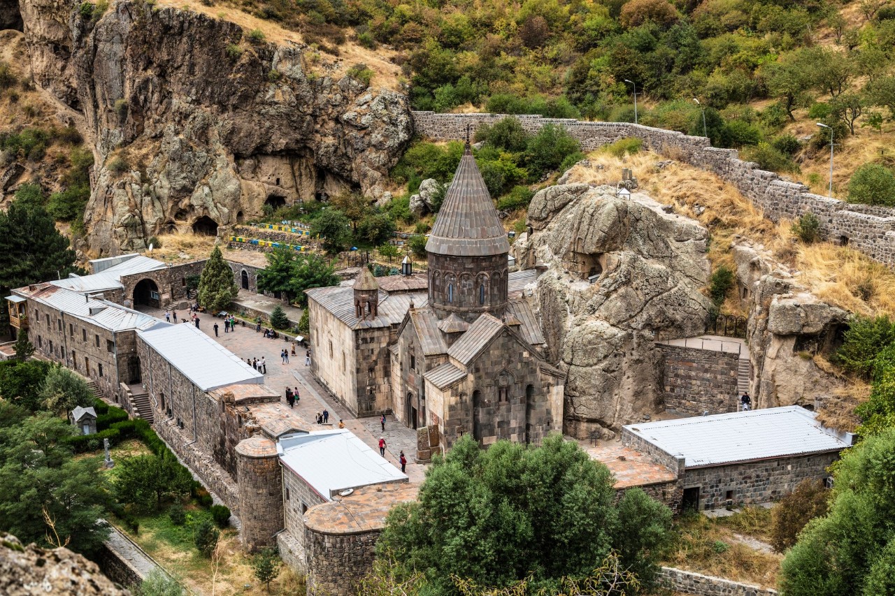 View from above of a large, grey monastery complex with a round tower and several outbuildings. The complex is surrounded by a wall and stands on a rock overgrown with bushes and trees.