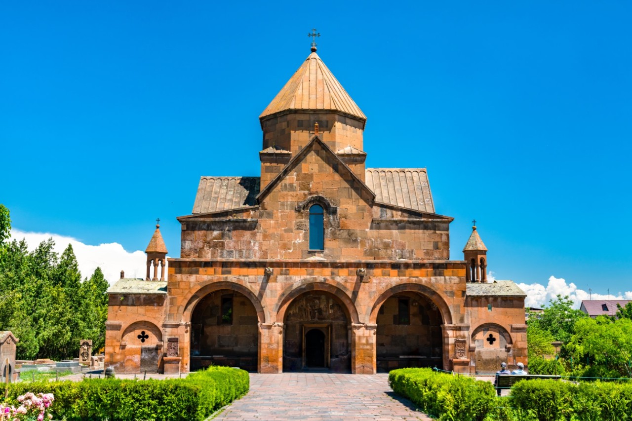 View of a three-nave basilica with a main dome in a green garden. Three arches can be seen in the entrance area.