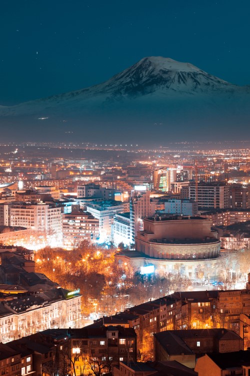 : View from above of a brightly lit city at night with snow-covered mountains in the background and a dark blue night sky.