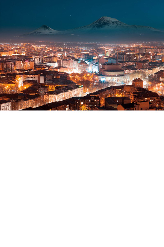 : View from above of a brightly lit city at night with snow-covered mountains in the background and a dark blue night sky.