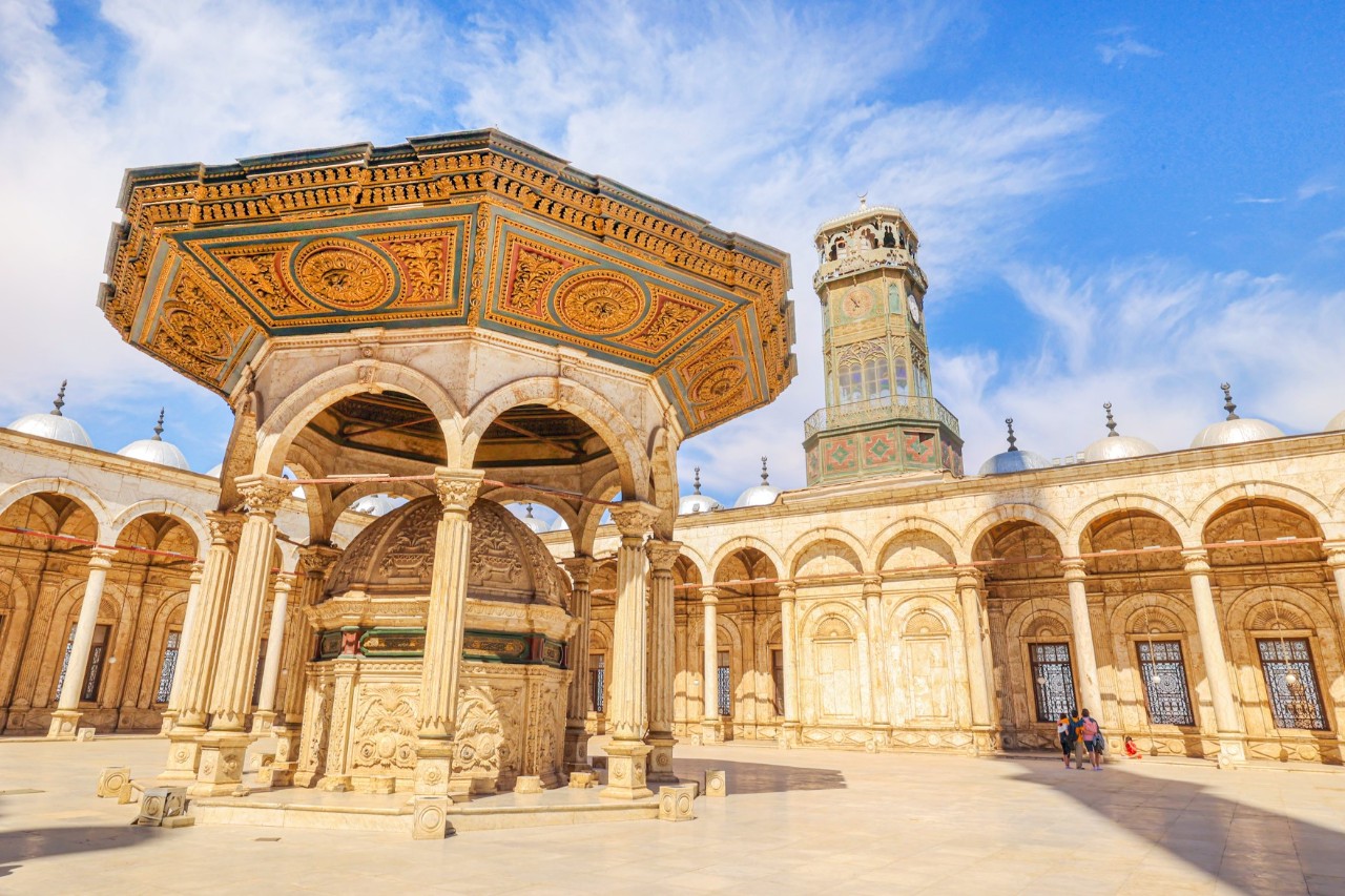 Interior courtyard of the Muhammad Ali Mosque in Cairo with an ornate ablution fountain supported by columns and topped with a richly decorated octagonal roof. In the background, a tall clock tower and a series of arched colonnades are visible under a partly cloudy sky.