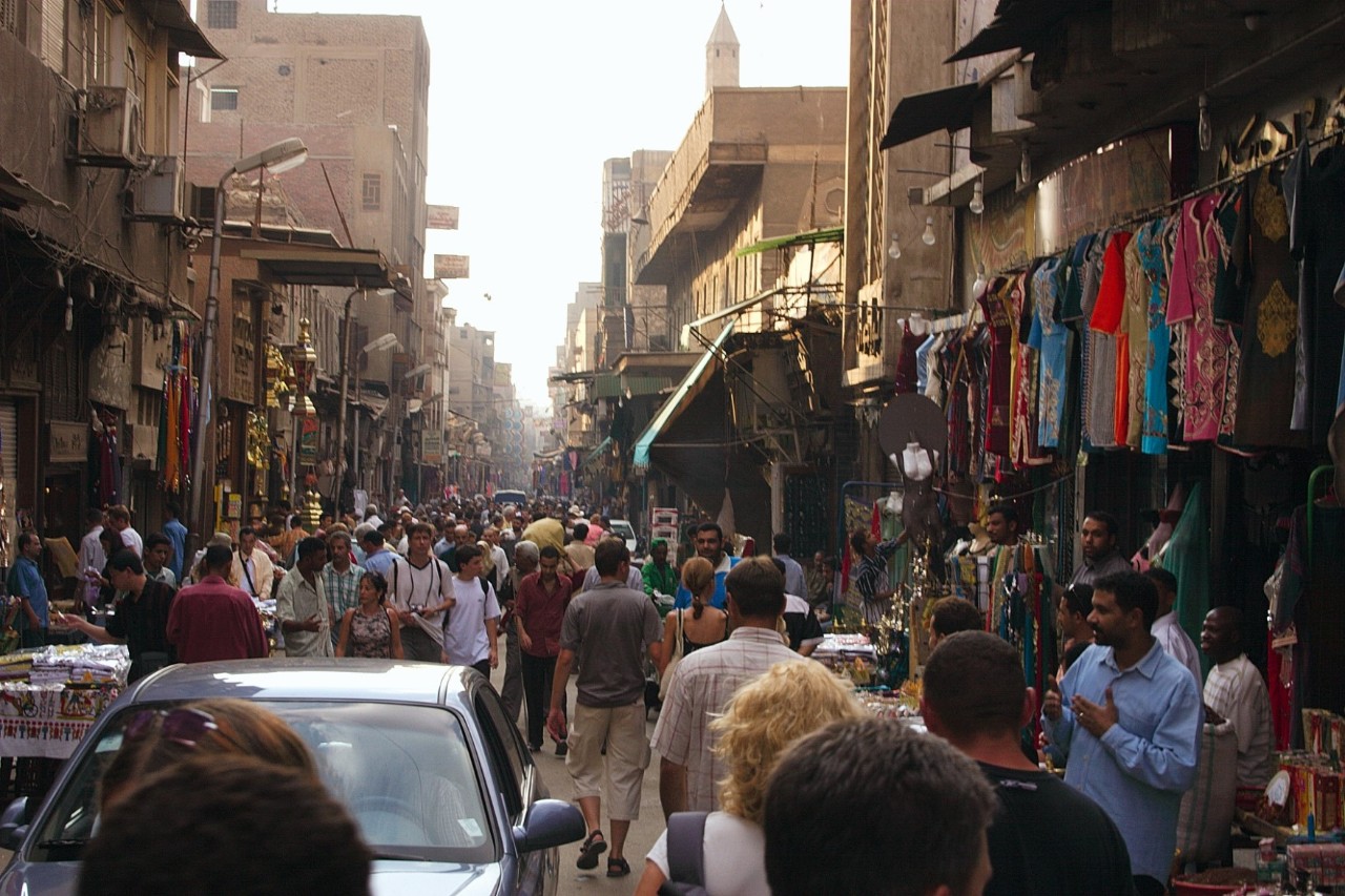 Busy market street in Cairo with many people, stalls, and colorful textiles hanging on building facades. Cars and pedestrians share the narrow alley between old buildings.
