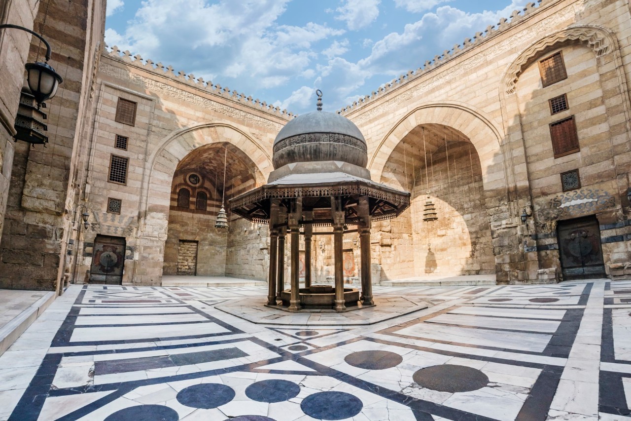 Courtyard of a historic mosque in Cairo with a central covered fountain on columns. The floor is decorated with geometric patterns in black and white marble; walls are made of light-colored stone.