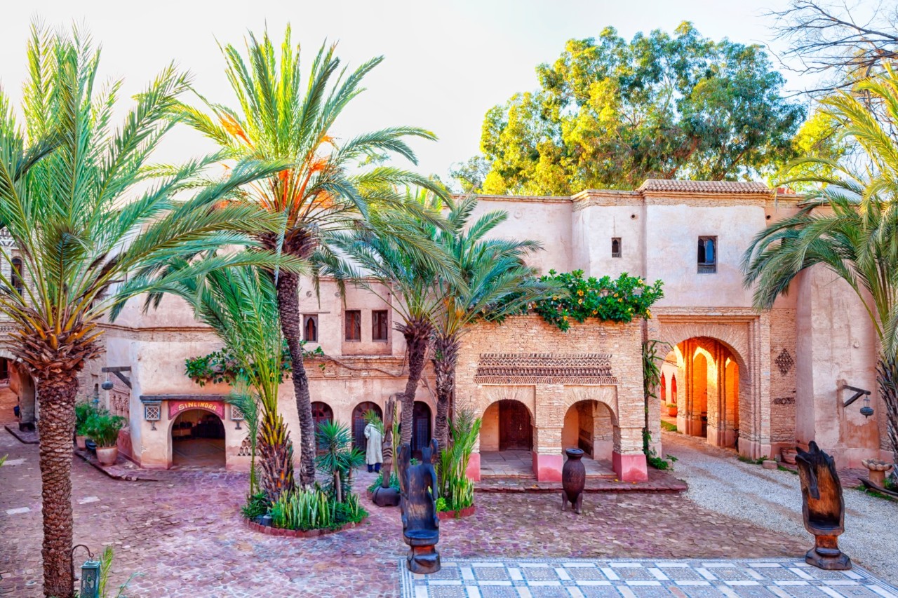 View of a two-storey, sand-coloured building with several arched entrances. A narrow alley leads through the largest arch of the building. In front of the building is a mosaic-tiled square with several palm trees and small wooden sculptures.