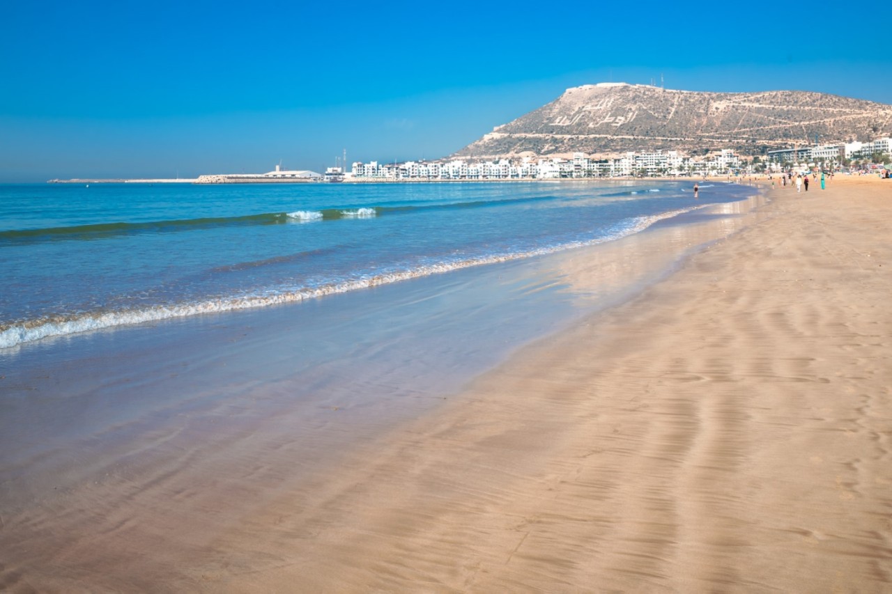 A light-coloured sandy beach stretches along the right-hand side of the picture, while the turquoise sea with gentle waves can be seen on the left. In the background, a large town with light-coloured houses can be seen in front of an imposing mountain.
