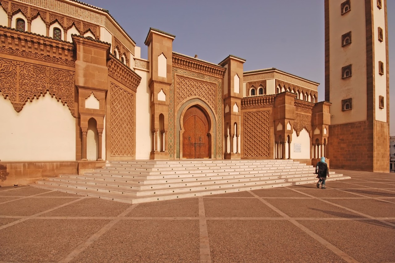 View of a reddish ochre-coloured building, with a wide staircase and a few short steps leading up to the entrance gate. The building’s facade is decorated with mosaics and stucco work. A person walks at the bottom of the steps. A tall, square, tower rises on the right-hand side of the building, the spire protruding from the picture. There are square windows on each floor on both visible sides of the tower.
