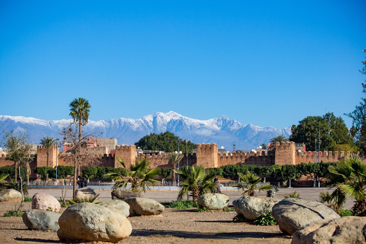 A fortress wall stretches across the picture from left to right. Treetops and the upper parts of buildings are visible behind the wall. A majestic mountain massif with snow-capped peaks rises in the background. In front of the wall is a square with scattered boulders and scattered low palm trees.