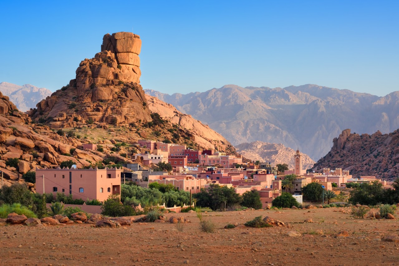 A small village with reddish houses below a huge, reddish rock formation that rises up into a tower-like shape. A mountain massif can be seen in the background.
